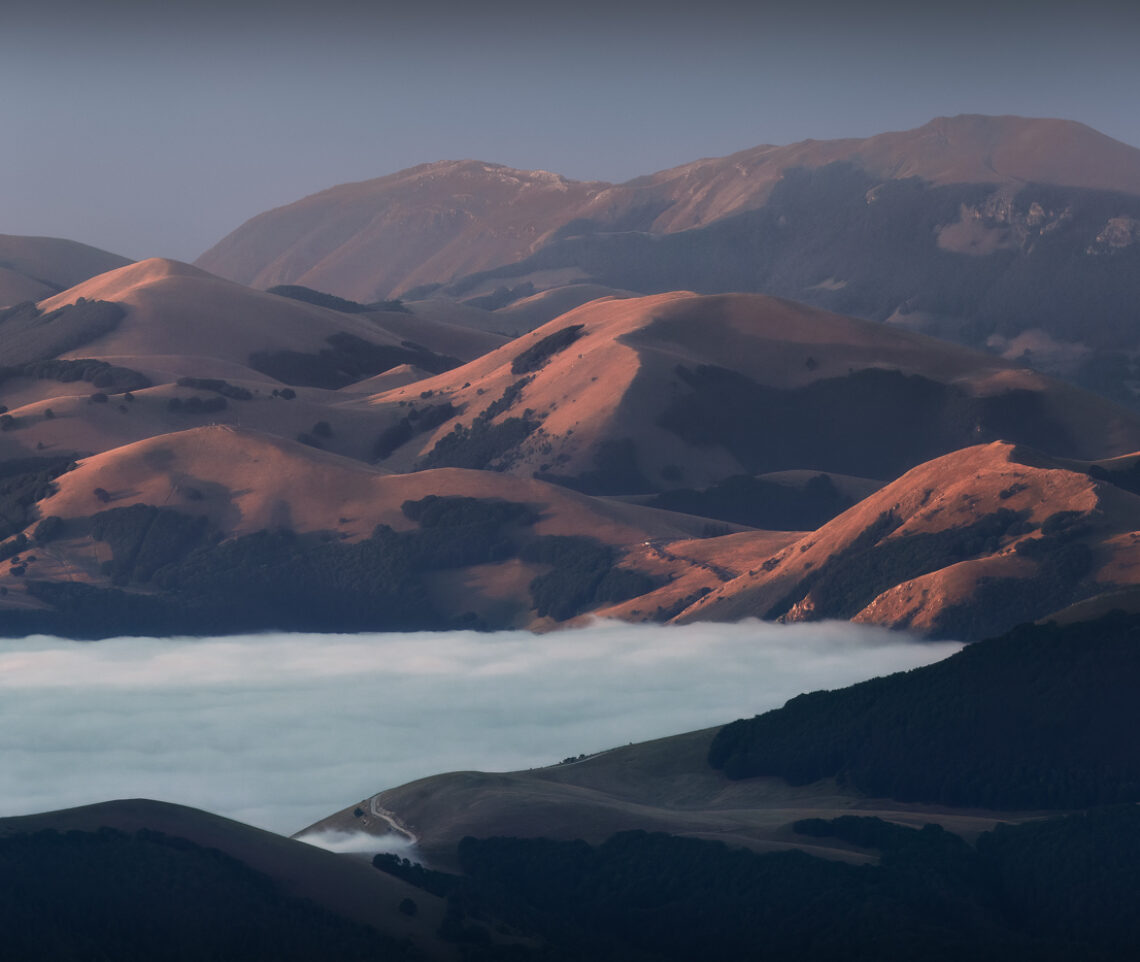 L’alba incendia i sinuosi versanti dei rilievi che emergono dalle nubi, ancora adagiate sulla grande piana di Castelluccio di Norcia. Parco Nazionale dei Monti Sibillini, Italia.