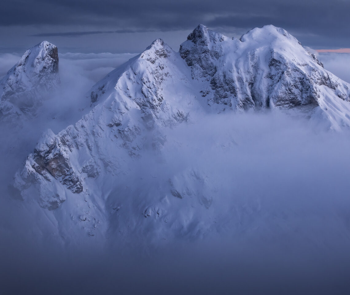 Cime sagomate dallo scenario invernale emergono da un mare di nubi per diventare anch’esse protagoniste dell’ora blu. Dolomiti Ampezzane, Italia.