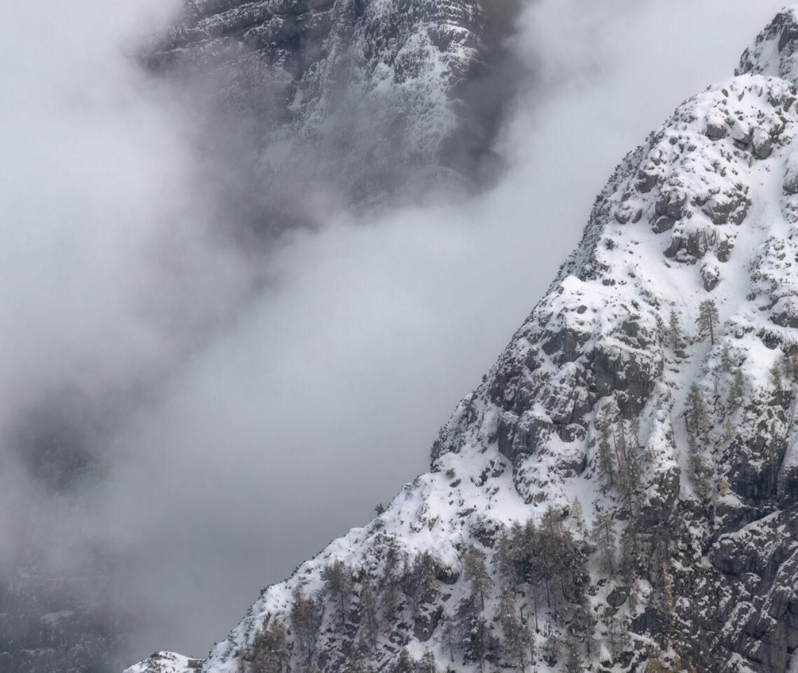 Colossali titani di roccia fanno breccia tra le nubi di una fredda giornata tardo autunnale. I larici si mettono al riparo come possono. Alpi Giulie, Italia.