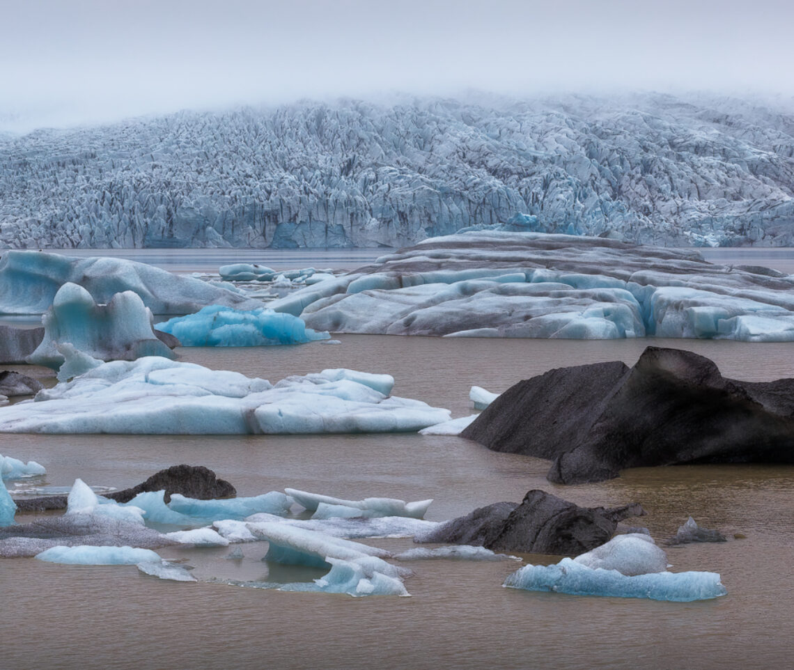 Il ghiacciaio Fjallsjökull fa breccia nelle nubi basse mostrando tutta la sua magnificenza, mentre il lago proglaciale formato da esso accoglie i suoi iceberg. Vatnajökull National Park, Islanda.