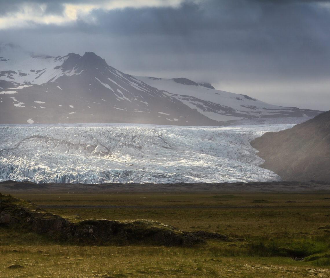 I raggi solari fratturano il cielo plumbeo di una giornata piovosa. Brilla sotto di esso il ghiacciaio Fláajökull. Vatnajökull National Park, Islanda.