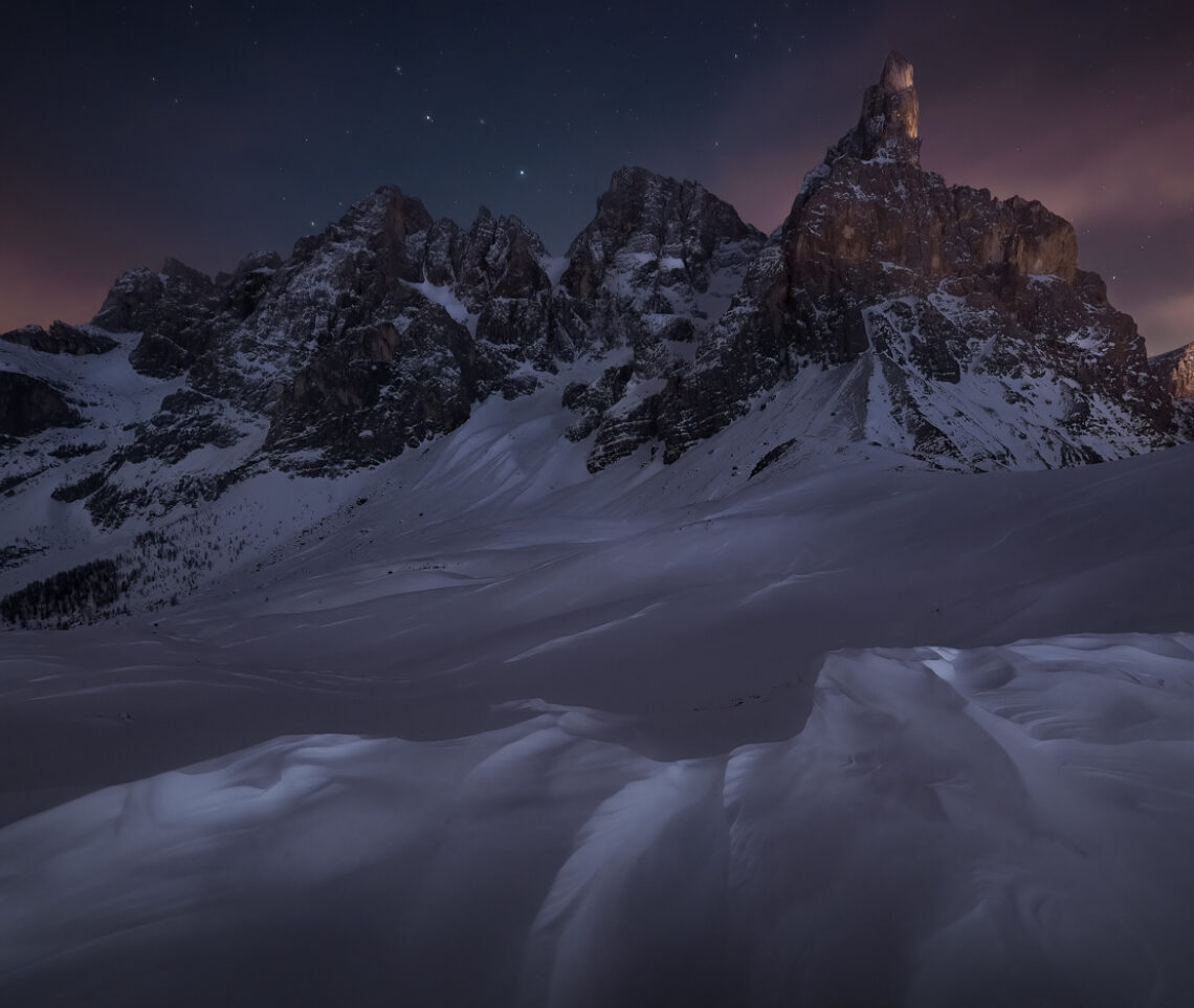 Il vento ha scolpito la superficie bianca che riveste i pendii delle Pale di San Martino. Una larga fenditura nel cielo notturno apre il sipario dell’infinito. Parco Naturale Paneveggio Pale di San Martino, Italia.